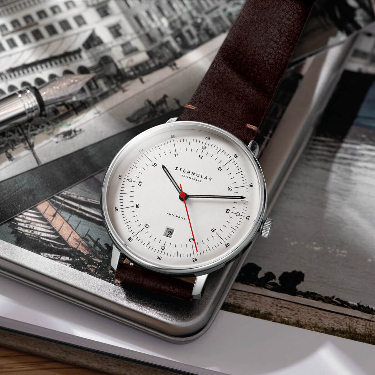ZEIT-Sonderedition Uhr »Hamburg« mit silbernem Gehäuse und braunem Lederarmband, roter Sekundenzeiger, minimalistisches Design, liegend auf einem Buch und einem Schwarz-Weiß-Foto.
