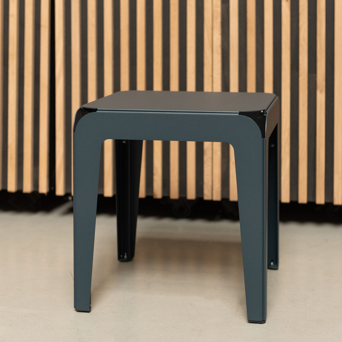 Ein moderner, minimalistischer Hocker namens »Bended Stool« mit schwarzer Sitzfläche und grauen Beinen vor einem Hintergrund in Holzoptik.