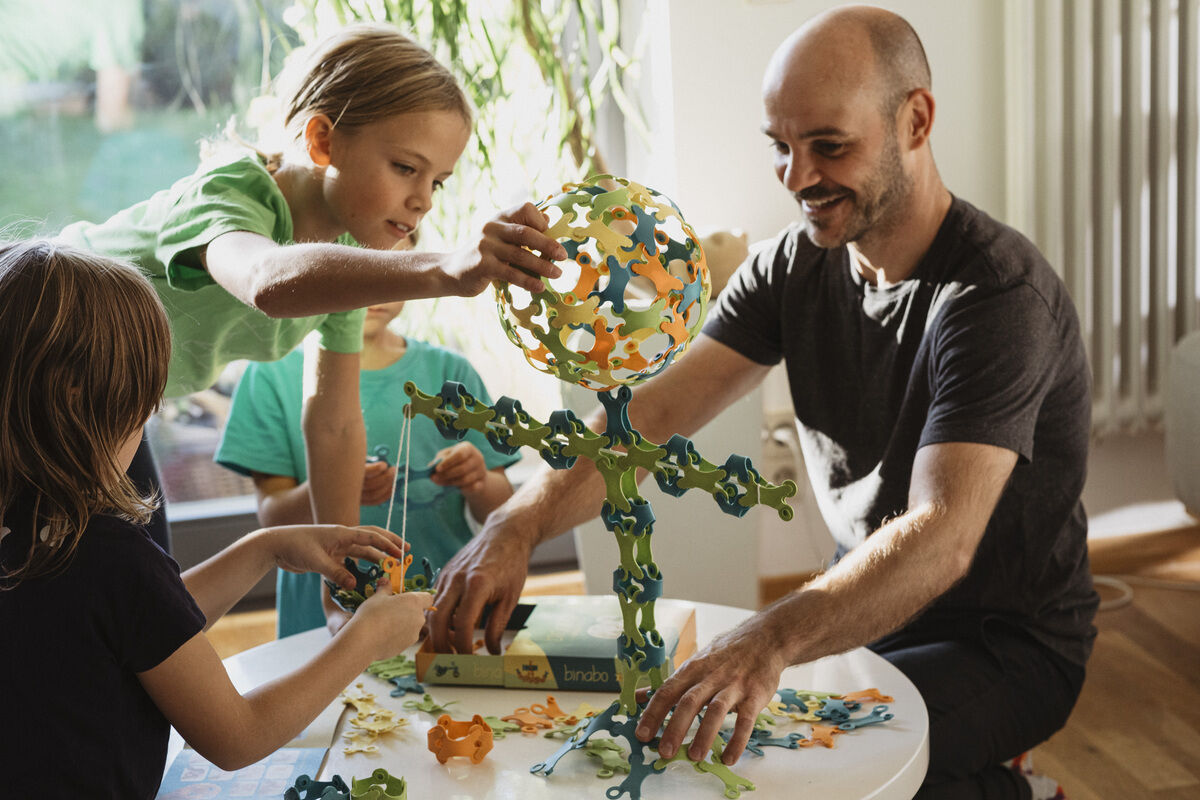 Ein Mann und Kinder sitzen gemeinsam an einem Tisch und spielen mit Binabo, einem Kreativspielzeug aus bunten, zusammensteckbaren Teilen. Holzspielzeug steht auf dem Tisch.