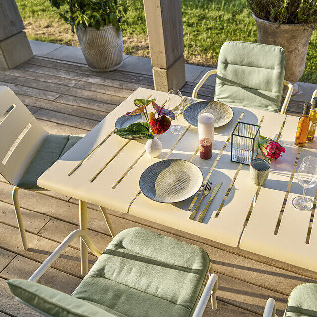 Ein schön gedeckter Tisch der Marke VAPIO (180x90 cm) auf einer Terrasse, umgeben von grünen Stühlen und Dekorationen. Die Szene ist in warmem Sonnenlicht, mit grünem Rasen im Hintergrund. Perfekt für Outdoor-Essen und Versammlungen.