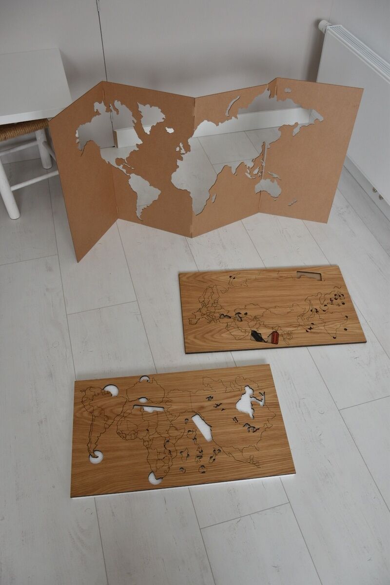 Eine selbstklebende Weltkarte aus Holz, bestehend aus drei Teilstücken unterschiedlicher Größe, auf einem weißen Holzboden liegend. Die Elemente der Karte zeigen verschiedene Kontinente und Inseln, zusammengefügt als dekoratives Wandbild.