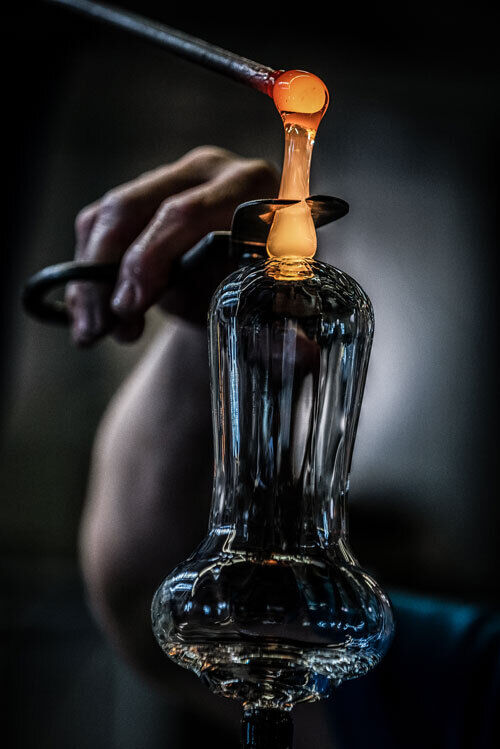 Handwerkliche Glasbläserei: Eine Hand formt ein orange glühendes Glas, das zu einem eleganten Sektglas verarbeitet wird, im Fokus.