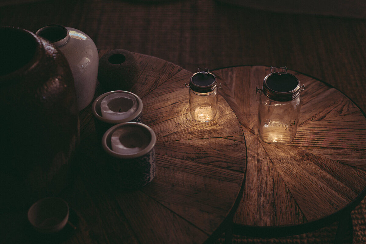 Zwei Sonnenglas-Glasbehälter mit integrierten LED-Solarlichtern, die auf einem hölzernen Tisch in einem dunklen, gemütlichen Raum stehen. Die Lichter erzeugen eine warme und stimmungsvolle Atmosphäre.