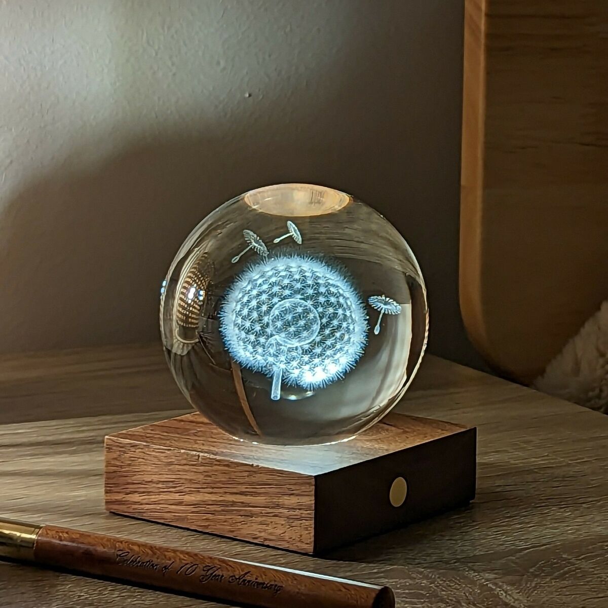 Auf einem Nachttisch steht die stilvolle Lampe »Amber Light« von Gingko Design Ltd mit einer runden Kristallglas-Kugel, die eine filigrane, leuchtende Pusteblume zeigt. Der Sockel besteht aus edlem Walnussholz, daneben liegt ein goldener Stift. Im Hintergrund ist ein Kissen sichtbar.