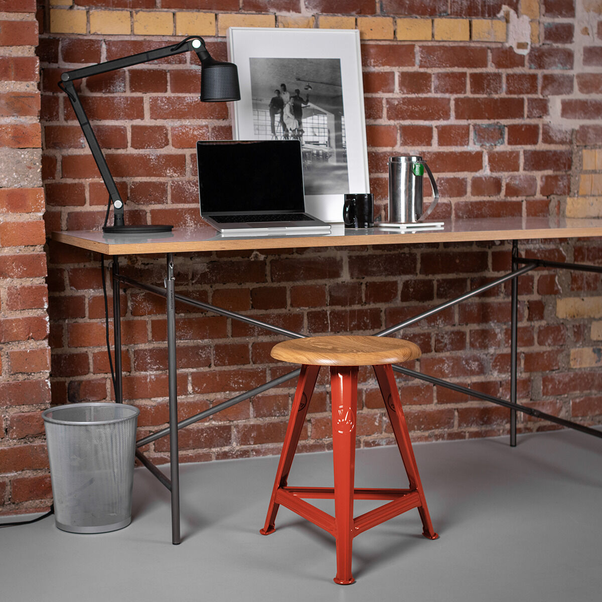 Moderner Schreibtischbereich mit einer industriellen Backsteinwand im Hintergrund. Auf dem roten Rowac-Schemel ist ein Holzsitz befestigt. Der Tisch hat eine Metallkonstruktion mit einer hölzernen Arbeitsplatte. Eine schwarze Lampe, ein Laptop und ein gerahmtes Bild stehen auf dem Tisch, begleitet von einem Becher und einer Thermoskanne.