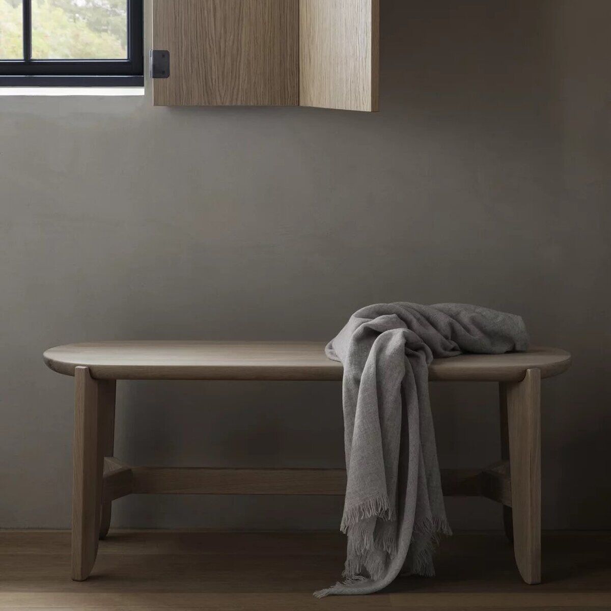 Sitzbank aus hellem Holz im minimalistischen Stil steht an einer Wand unter einem Fenster. Auf der Sitzfläche liegt eine gefaltete, graue Decke. Die Umgebung wirkt modern und ruhig, mit Akzent auf natürlichen Materialien.