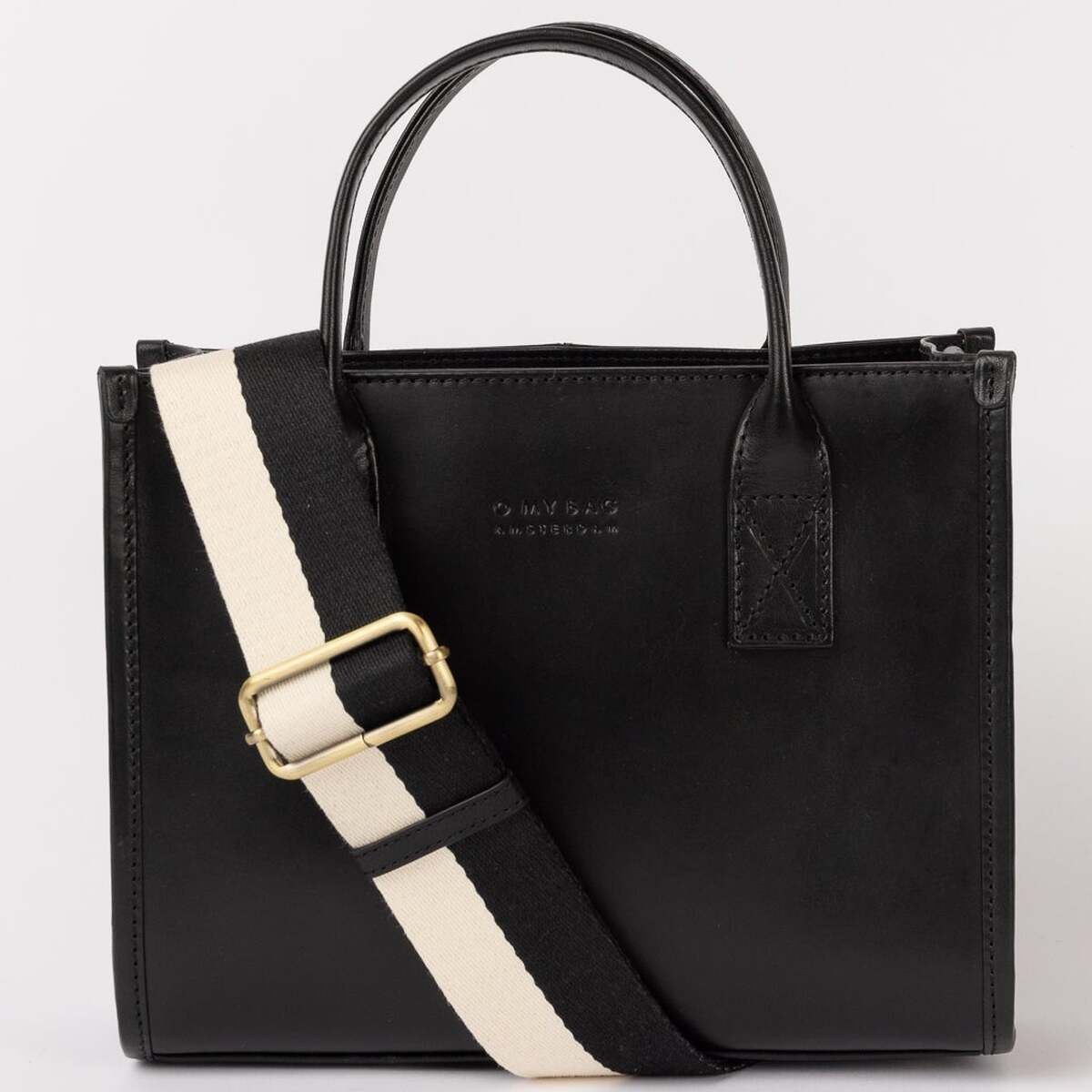 Schwarze Ledertasche mit kurzem Henkel und einem abnehmbaren cremeweißen Schulterriemen mit goldener Schnalle, stehend vor neutralem Hintergrund.
