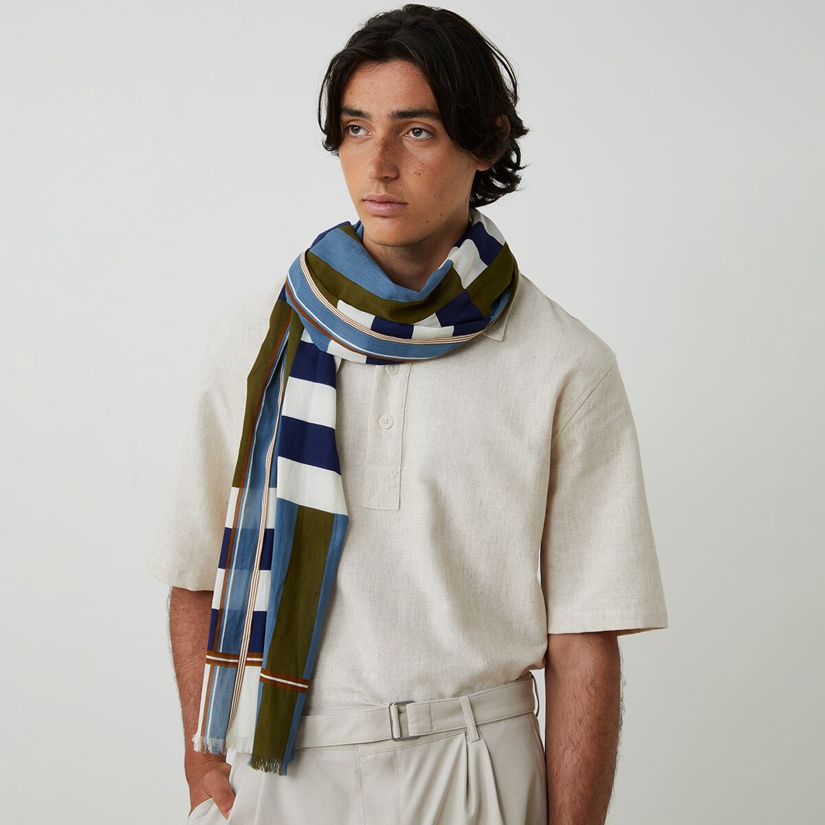Ein Mann trägt einen Baumwollschal No. 771 von MOISMONT in Blau, mit weißen, olivgrünen und dunkelblauen Streifen. Das Outfit besteht aus einem beigen, kurzärmeligen Hemd und einer hellen Hose. Der Schal wirkt leicht, modern und ist um den Hals geschlungen.