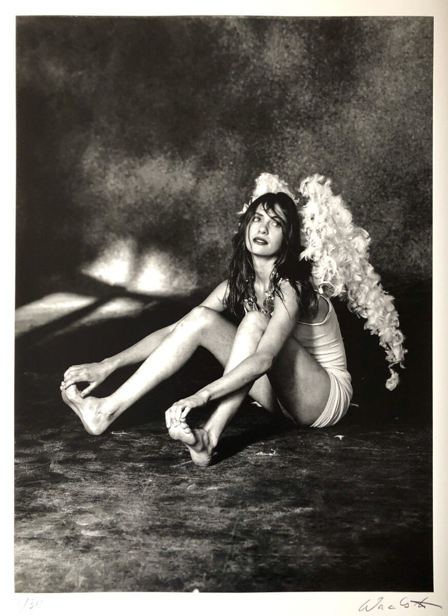 Eine Schwarz-Weiß-Fotografie zeigt eine Frau in sinnlicher Pose mit Engelsflügeln, sitzend auf dem Boden, umgeben von Licht- und Schatteneffekten. Es ist ein künstlerisches Werk mit mystischem Ausdruck, signiert von Wackerbarth.
