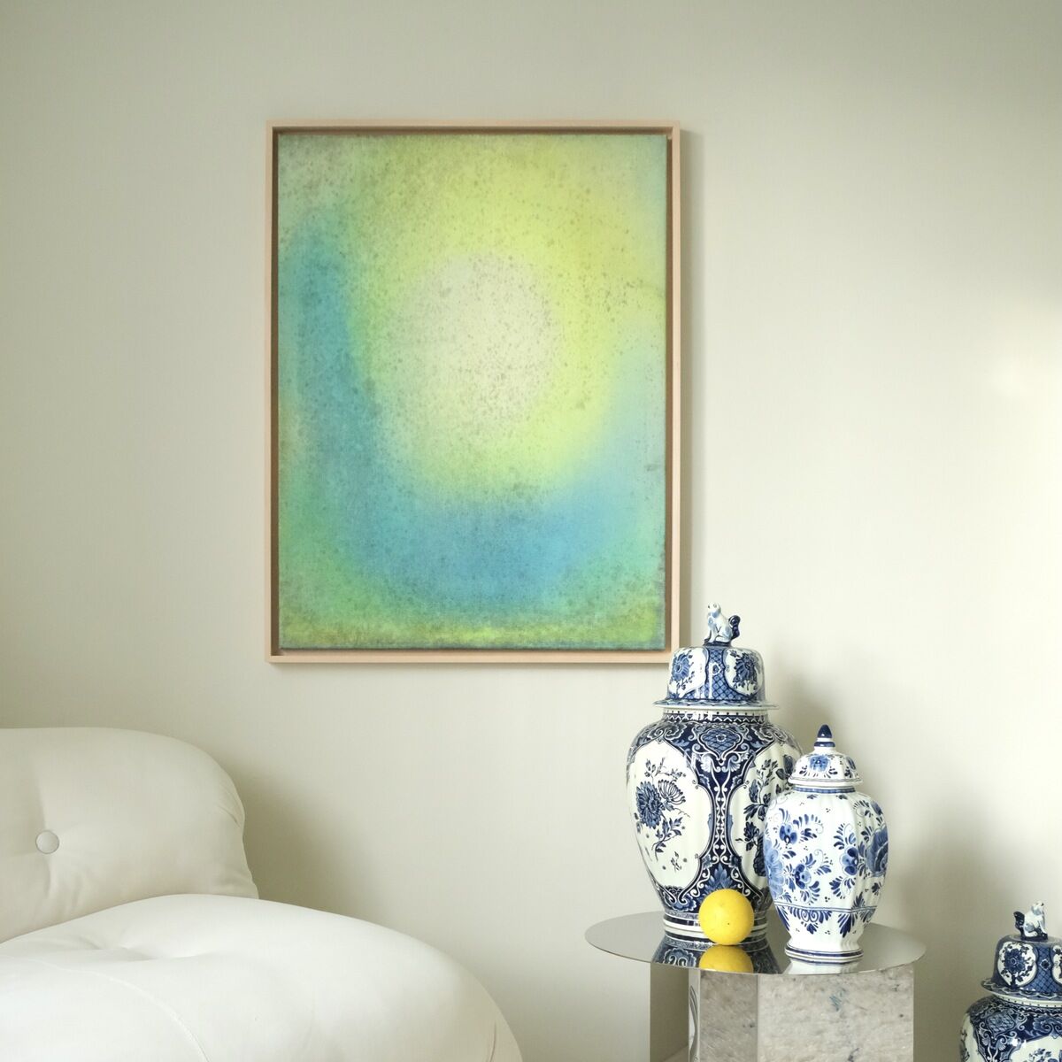 Ein gerahmtes abstraktes Kunstwerk in Blau-, Grün- und Gelbtönen hängt an der Wand eines modernen Wohnzimmers. Neben einer cremefarbenen Couch steht ein kleiner Tisch mit blauen und weißen Porzellanvasen.