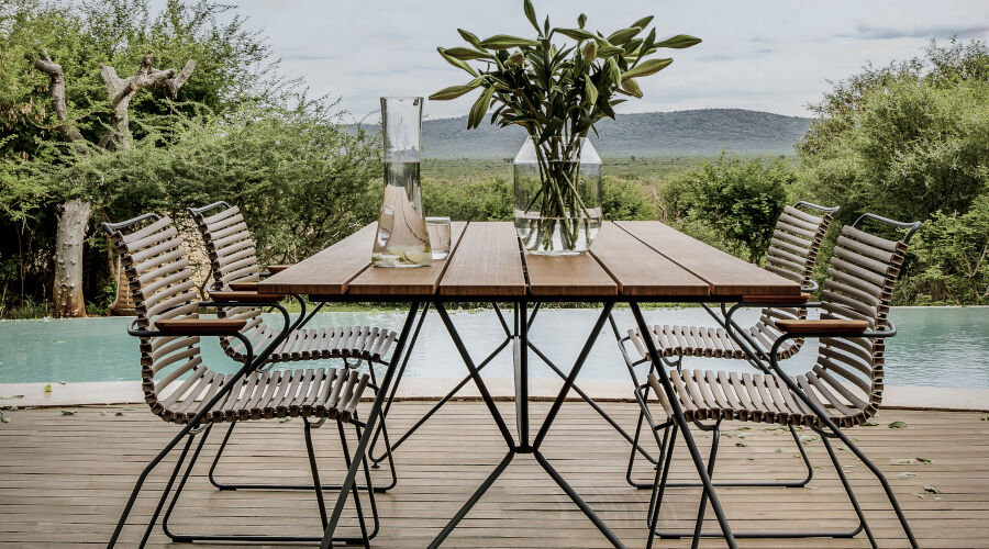 Moderner Gartentisch aus Holz mit Metallgestell, umgeben von sechs Stühlen mit Metallrahmen und Sitzflächen aus Bändern, auf Holzterrasse vor Pool und grüner Landschaft