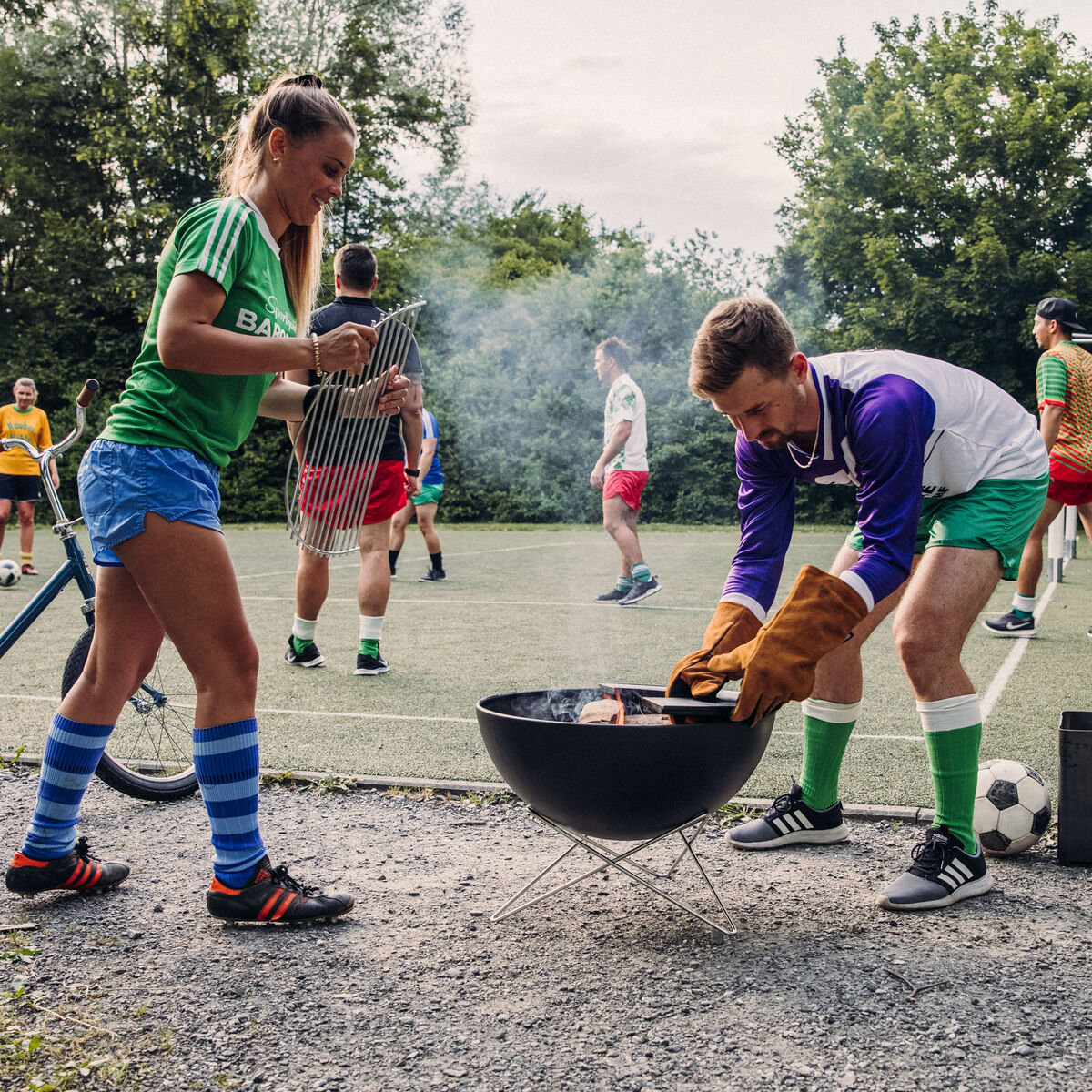 Eine Gruppe von Menschen spielt Fußball im Hintergrund, während zwei Personen die Feuerschale »BOWL 57« benutzen. Die Szene findet auf einem Outdoor-Sportplatz statt; das Wetter ist sonnig und die Umgebung ist grün und einladend. Fahrrad und Sportutensilien sind im Bild ebenfalls erkennbar.