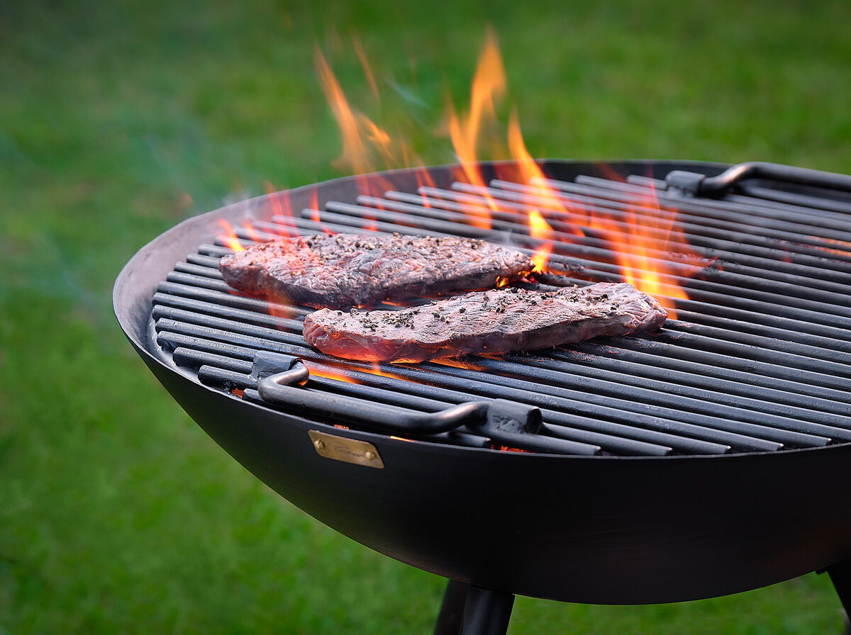 Runder Grillrost mit brennenden Flammen und zwei Steaks, die auf dem Grillrost garen. Scharfer Rasen im Hintergund.