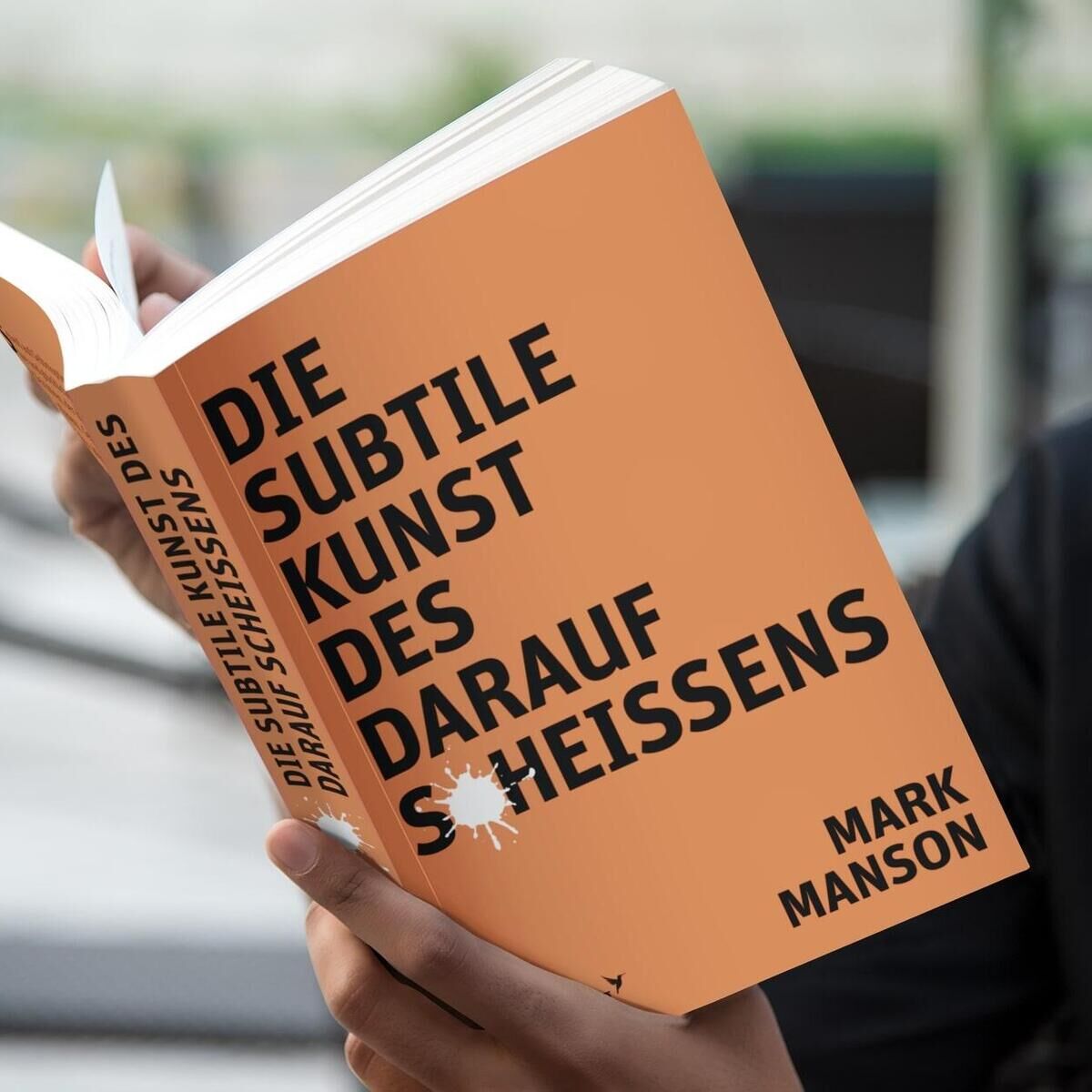 Eine Person hält das Buch 'Die subtile Kunst des darauf Scheißens' von Mark Manson in den Händen. Das Cover des Buches ist orange mit schwarzer und weißer Schrift.