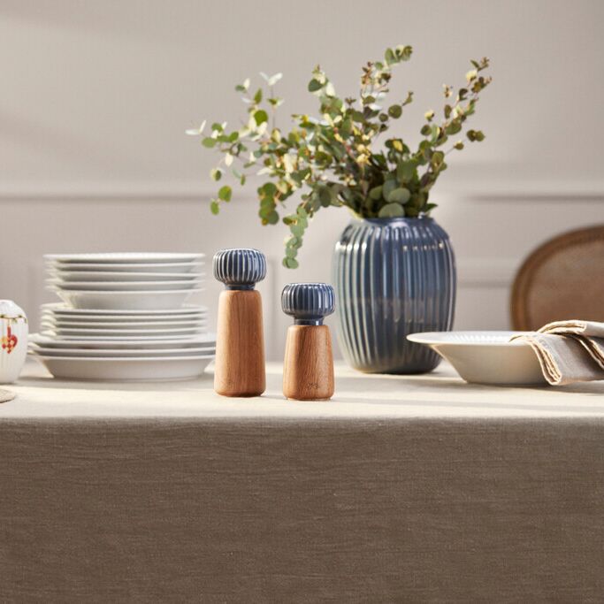 Zwei Gewürzmühlen des Modells Hammershøi aus Holz und Keramik stehen auf einem gedeckten Tisch mit hellgrauer Tischdecke, umgeben von weißen Tellern, einer blauen Vase mit Grünpflanzen und weiterem Geschirr in elegantem, modernen Stil.
