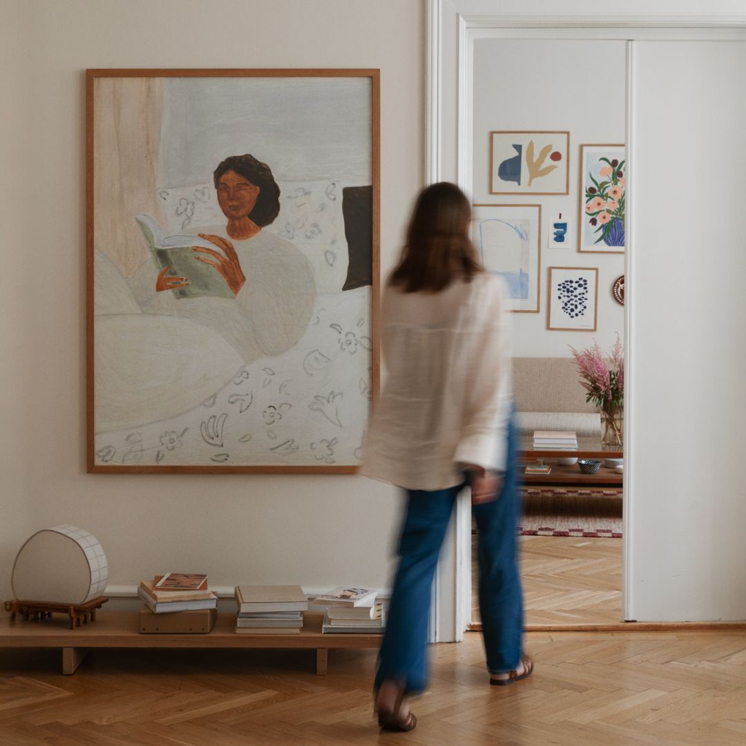 Ein moderner Wohnraum mit einem großen Gemälde an der Wand, das eine Person beim Lesen zeigt. Eine Frau im Vordergrund, ein Holzboden, ein Regal mit Büchern und eine helle, minimalistische Einrichtung.