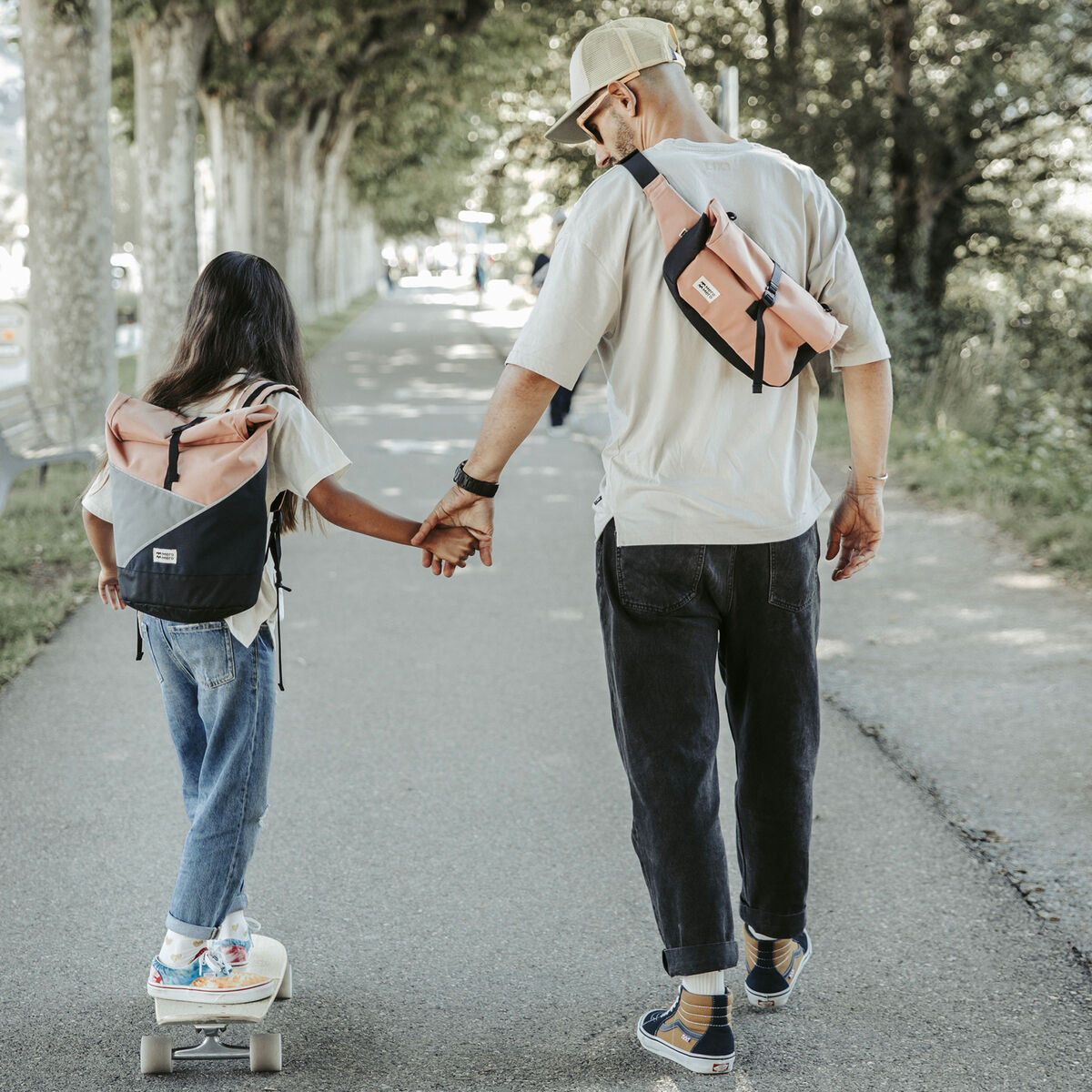 Ein Erwachsener und ein Kind sind von hinten zu sehen, wie sie Hand in Hand auf einem Waldweg spazieren gehen. Beide tragen beige-rosa Taschen der Marke 'Popoyo'. Das Kind fährt auf einem Skateboard und trägt eine Jeans und ein graues T-Shirt. Der Erwachsene trägt schwarze Hosen, eine beige Kappe und eine weiße Jacke.