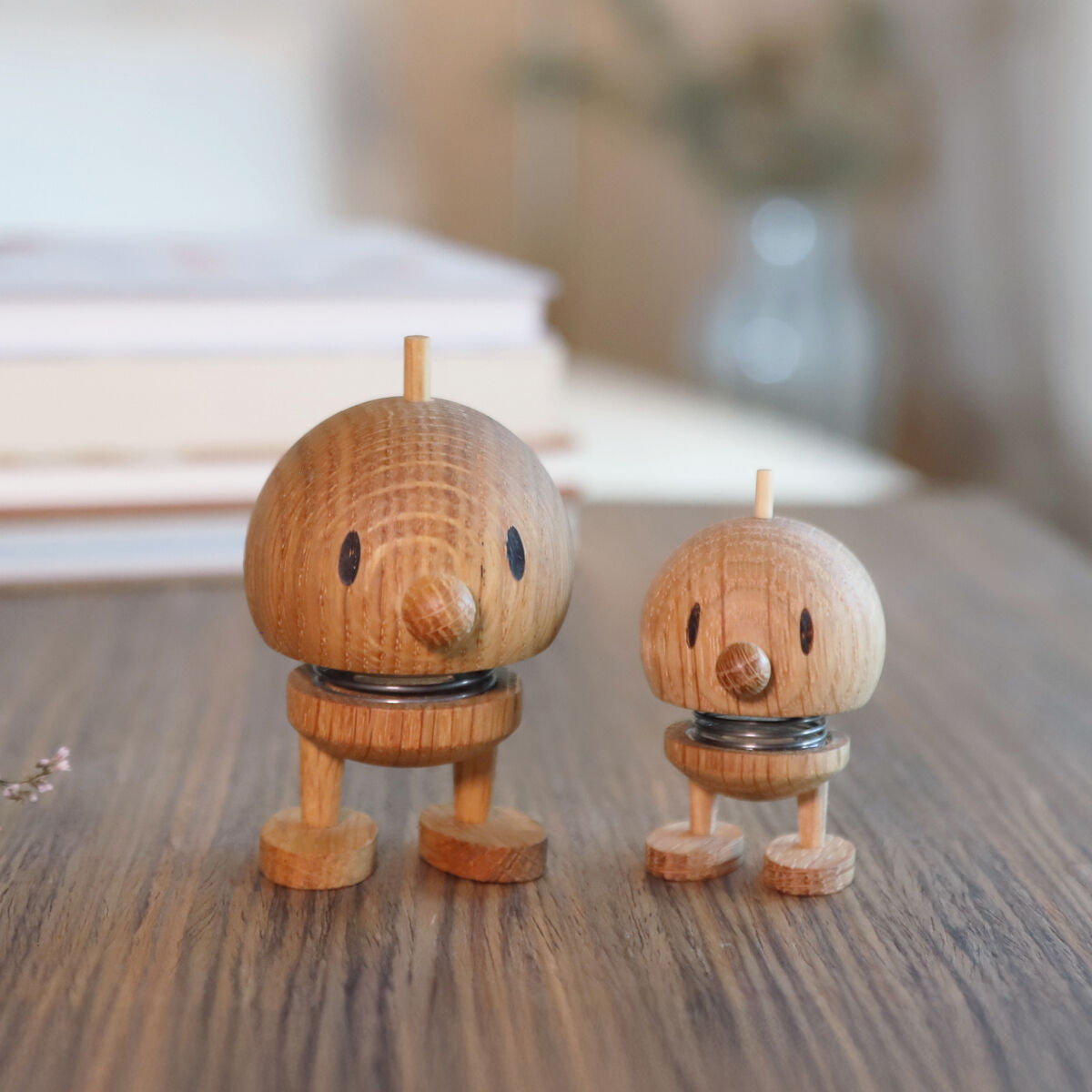 Zwei hölzerne Hoptimisten in verschiedenen Größen aus Eiche stehen auf einem Tisch vor einem hellen, unscharfen Hintergrund mit einem Bücherstapel. Die Figuren wirken freundlich und dekorativ, ideal als Geschenk oder Dekoration für moderne Wohnräume.