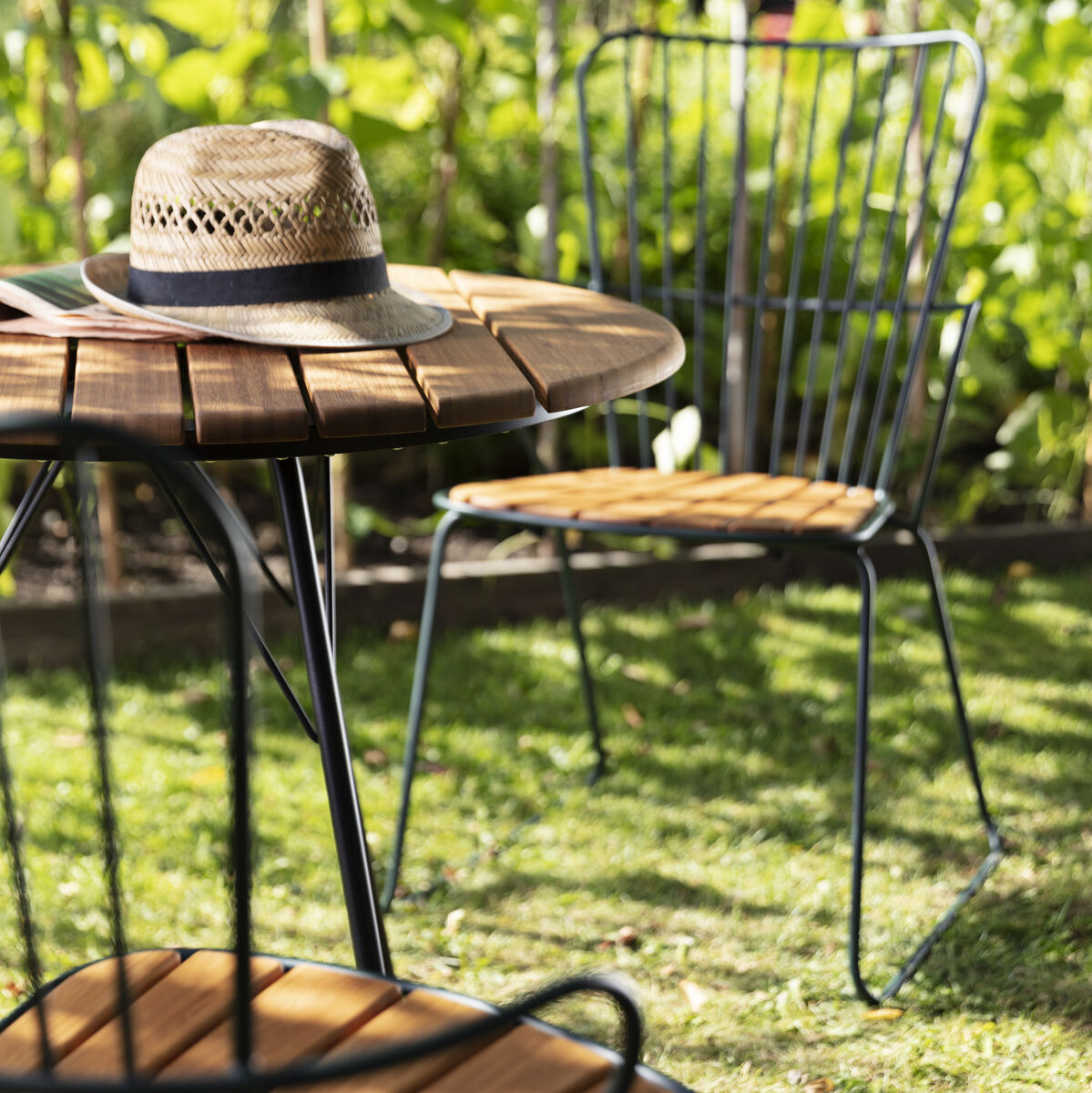 Zwei PAON Essstühle aus Stahlrahmen mit Holzsitzflächen stehen im Grünen neben einem runden Holztisch. Einer der Stühle hat einen Sonnenhut auf dem Sitz.