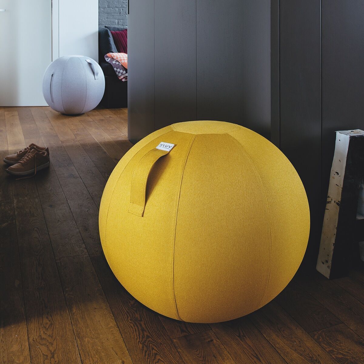 Ein gelber Sitzball der Marke „Vluv Leiv“ steht in einem modern eingerichteten Wohnzimmer auf einem Holzboden. Im Hintergrund ist ein grau-weißer Sitzball und ein orange gemustertes Kissen auf einem grauen Sofa zu sehen.