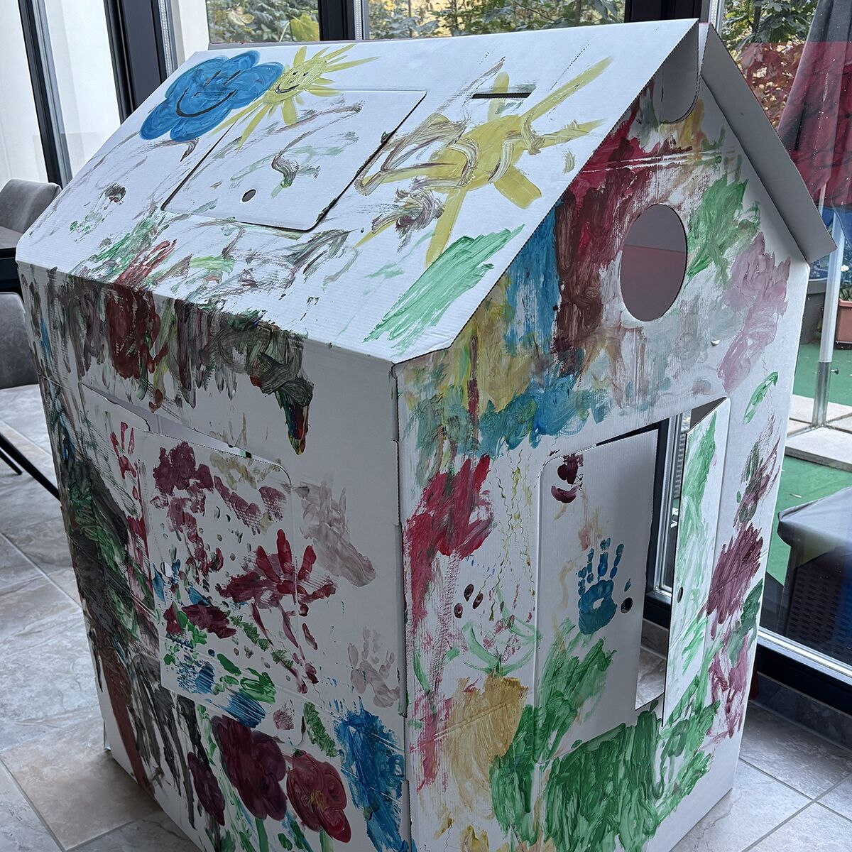 Ein großes, zusammengebautes Spielhaus aus stabiler weißer Wellpappe mit bunten, handgemalten Motiven und vielen Handabdrücken, steht in einem hellen Raum mit großen Fenstern. Das Dach zeigt Sonne, Wolken und Tiere, Wände sind mit Fingerfarben bemalt.