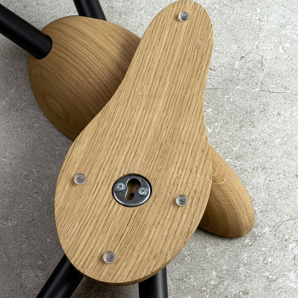 Detailaufnahme der Wanddeko »RESPECT« von finbeaux aus massiver Esche mit drei schwarzen Metallbeinen, auf hellem Steinboden liegend. Sichtbar sind außerdem eine Schraube und ein Dübel. Klare Holzmaserung und dezente Metallakzente, modernes Design.
