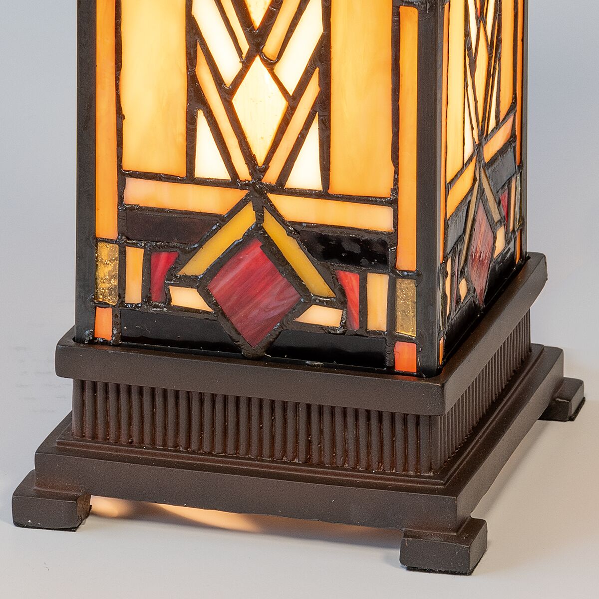Detailaufnahme des Sockels einer Tischleuchte im Tiffany-Stil mit geometrischen Mustern in Bernstein-, Rot-, Schwarz- und Beigetönen. Der Fuß ist dunkelbraun, gerippt und rechteckig. Das Licht scheint warm durch das farbige Glas, betont die kunstvolle Gestaltung.