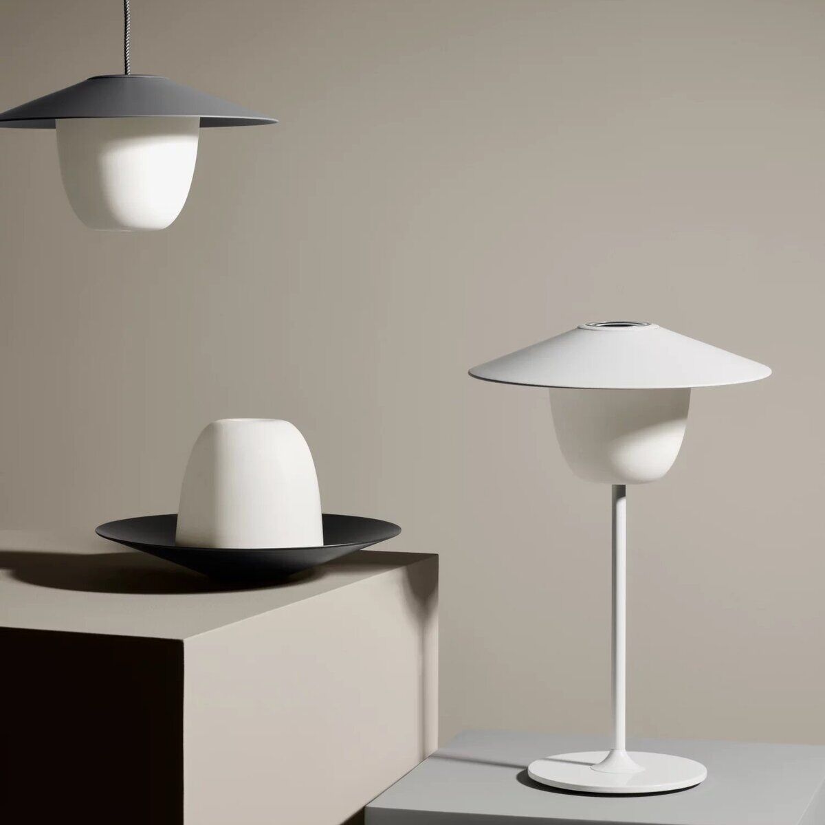 Die mobile LED-Tischleuchte »ANI« zeigt drei moderne, minimalistische Designs. Ein hängendes Modell hat einen weißen Schirm mit grauer Oberseite, während zwei stehende Modelle auf Podesten platziert sind. Eine Lampe ist kompakt und schwarz mit weißem Schirm, die andere hat einen schlanken weißen Fuß und breiten Schirm. Die Farben der Lampe sind weiß, schwarz und grau.