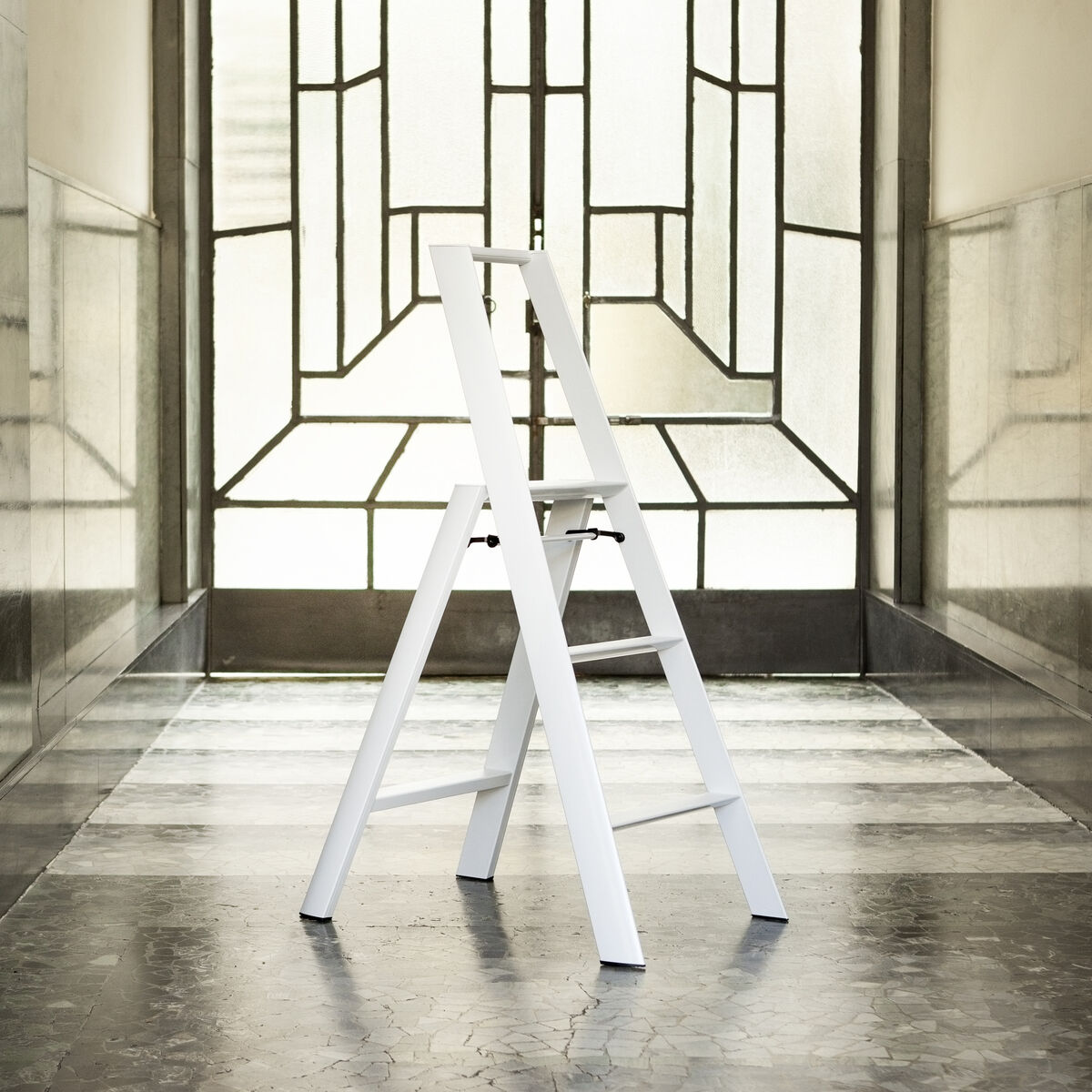 Eine weiße dreistufige Trittleiter steht in einem hellen, modern gestalteten Flur vor einem großen Fenster mit geometrischem Muster. Der Raum wirkt großzügig und minimalistisch, die Leiter ist zentral platziert und bereit zum Gebrauch.