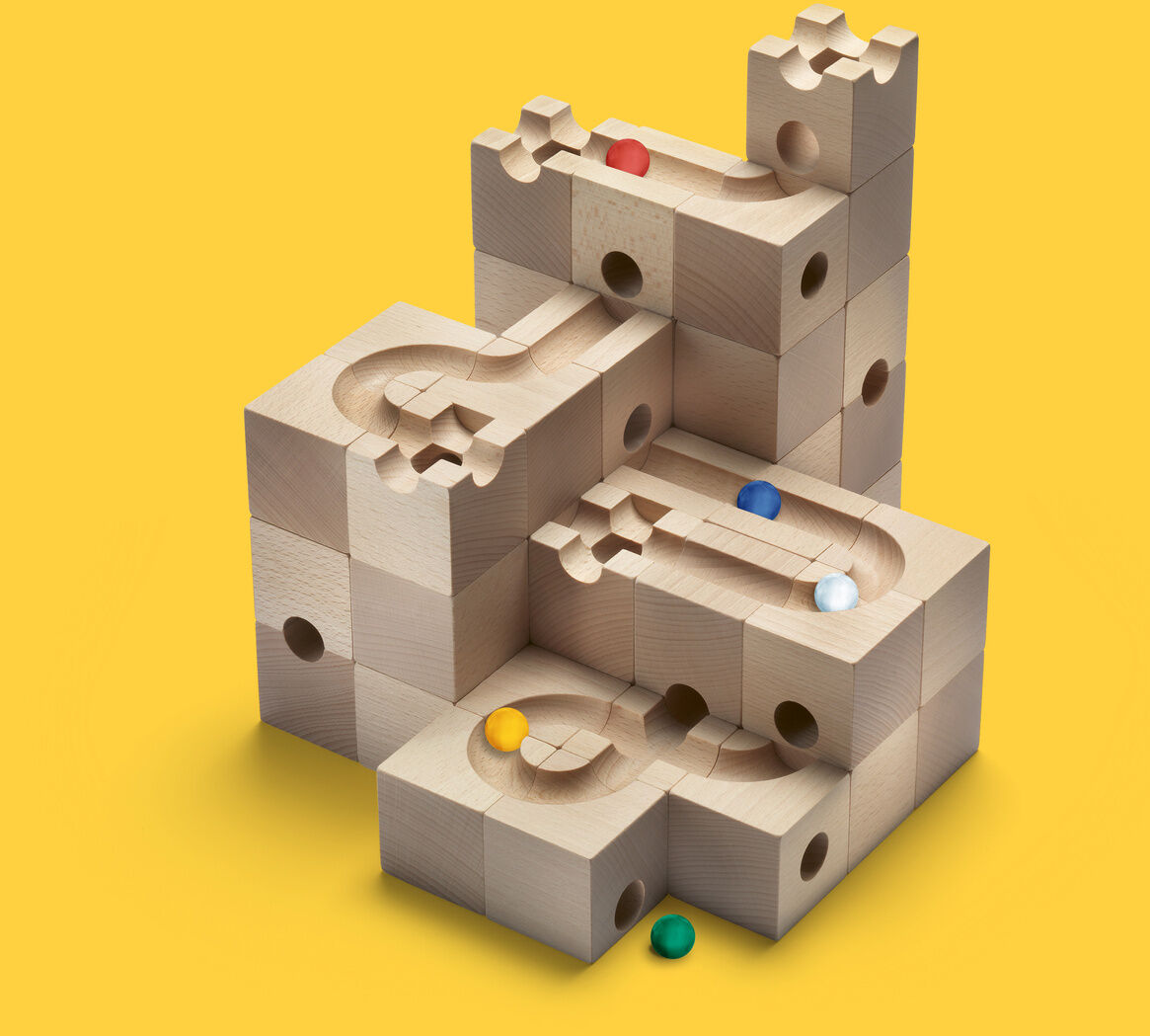 Das Bild zeigt das Kugelbahn Starter-Set »STANDARD« auf einem gelben Hintergrund. Es besteht aus hölzernen Bausteinen, die zu einer Murmelbahn zusammengebaut sind und enthält bunte Murmeln in verschiedenen Farben.