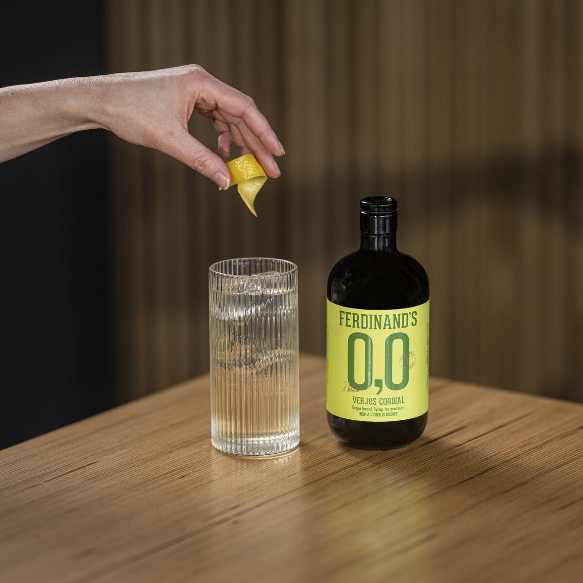 Eine Hand hält eine Zitronenzeste über ein mit Eis gefülltes Glas neben einer Flasche Verjus Cordial 0,0 von Ferdinands auf einem hellen Holztisch. Das Getränk sieht erfrischend und klar aus, die Szene vermittelt eine moderne, gesunde Atmosphäre.