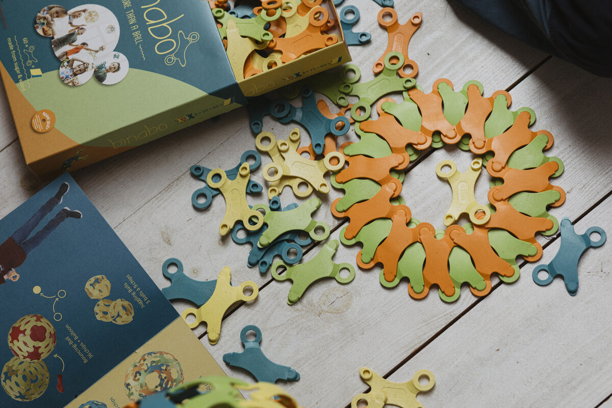Das Bild zeigt das Spiel »Binabo« mit 60 Teilen. Verschiedene grüne, gelbe, orange und blaue Steckteile wurden teilweise miteinander verbunden und liegen ausgebreitet auf einem hellen Holzboden. Die Verpackung des Spiels ist ebenfalls sichtbar.
