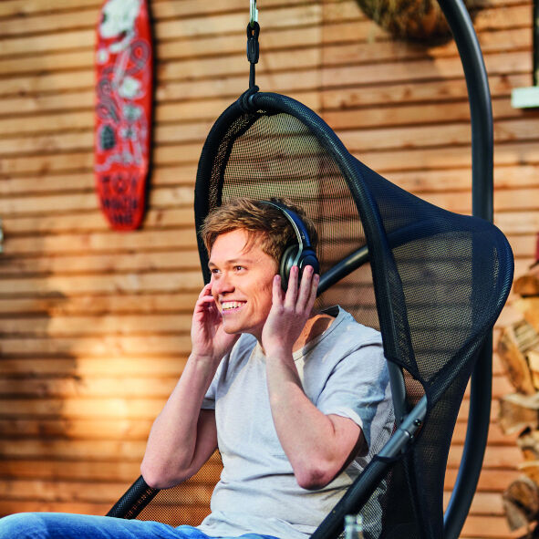 Ein junger Mann sitzt in einem schwarzen Hängesessel der Marke »Yobaya« und hört mit Kopfhörern Musik, vor einer Holzwand dekoriert mit verschiedenen Dekorationsgegenständen. Die Atmosphäre wirkt gemütlich und einladend.