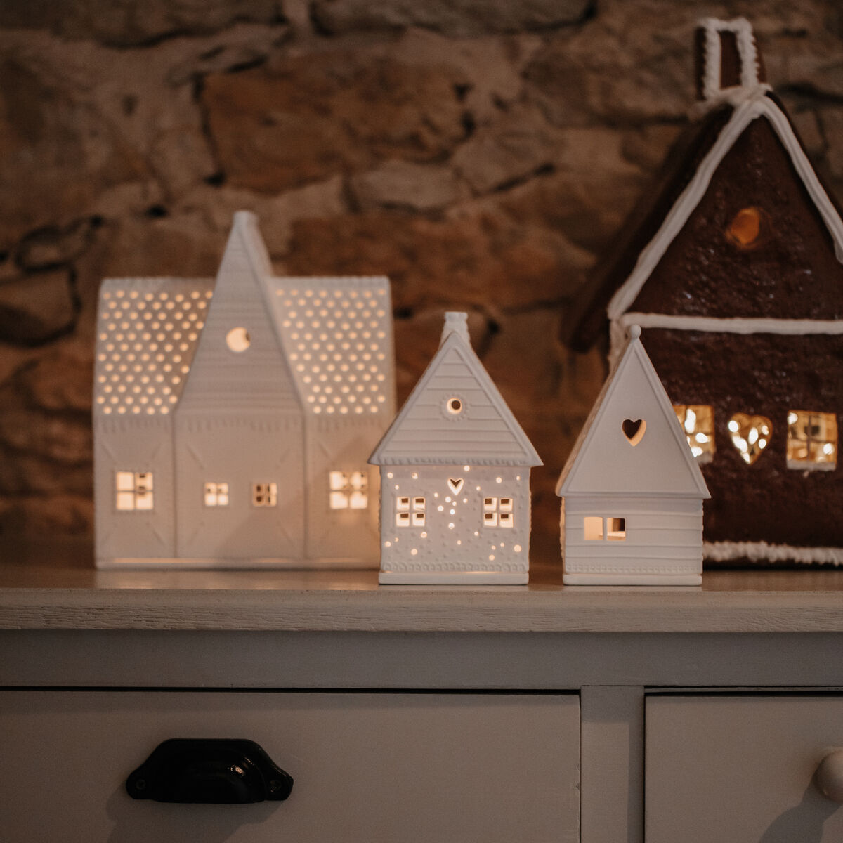 Drei Lebkuchen- und Lichthäuser stehen auf einem Tisch vor einer Steinwand, mit warmem Licht, das eine gemütliche und festliche Atmosphäre schafft.