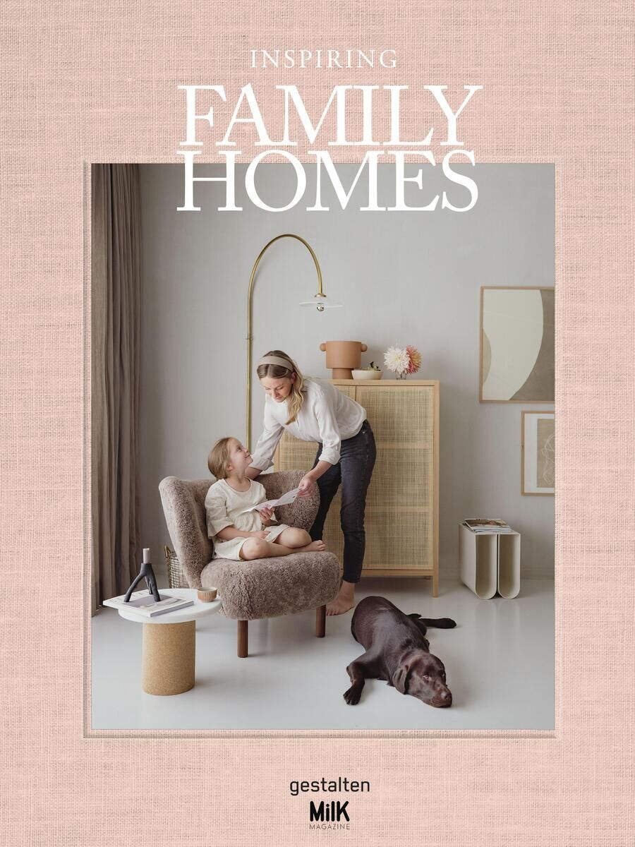 Ein Buchcover mit dem Titel 'Inspiring Family Homes' zeigt eine moderne, liebevolle Familienszene. Eine Frau beugt sich zu einem sitzenden Kind hinunter, während ein Hund auf dem Boden liegt. Das Zimmer ist modern und minimalistisch eingerichtet mit neutralen Farben.