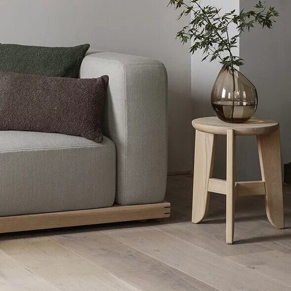Ein moderner, minimalistischer Sitzhocker aus hellem Holz steht neben einem hellen Sofa im Wohnzimmer. Auf dem Sofa liegen Kissen in Grün und Braun sowie eine beigefarbene Decke. Daneben steht eine Glasvase mit Zweigen auf dem Hocker. Der Raum ist in natürlichen, dezenten Farben gehalten.