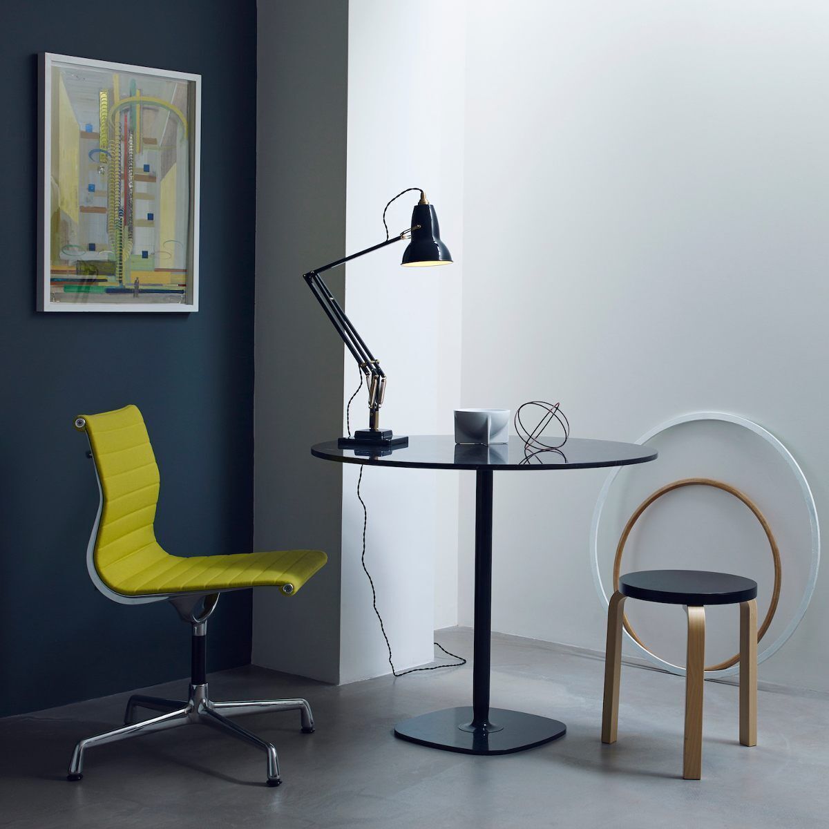 Eine moderne Tischleuchte »Anglepoise Original 1227 Brass Desk Lamp« steht auf einem runden, schwarzen Tisch. Im Hintergrund befinden sich ein gelber Bürostuhl, ein Kunstwerk an einer dunkelgrauen Wand und ein dekorativer Reif. Die Lampe hat ein schwarzes und messingfarbenes Design und ist in einer minimalistischen, stilvollen Umgebung platziert.
