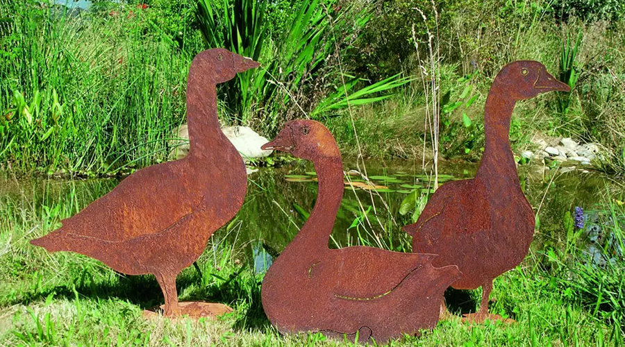 Drei rostige Metallskulpturen in Form von Gänsen stehen auf Gras vor einem Teich mit grüner Vegetation