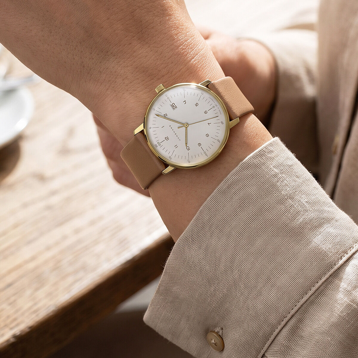 Die Junghans ZEIT-Sonderedition »max bill Damen« zeigt ein elegantes, rundes Edelstahlgehäuse mit sandgoldener, matter PVD-Beschichtung, weißem Zifferblatt, schmalen Indizes und hellbraunem Lederarmband am weiblichen Handgelenk. Dezenter, moderner Look.