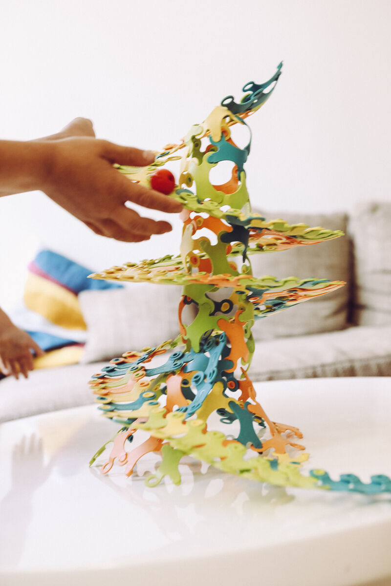 Ein bunter Turm aus ineinander steckbaren Kunststoffteilen des Spiels 'Binabo'. Eine Hand greift nach dem Turm. Im Hintergrund ist ein Sofa und Kissen zu sehen. Das Spielzeug besteht aus grünen, orangen, blauen und gelben Teilen.