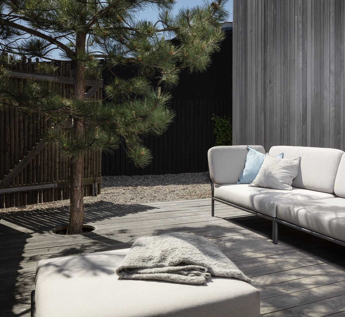 Eine moderne Lounge-Ecke auf einer Holzterrasse im Freien. Sie besteht aus einem grauen Ecksofa und einem passenden Ottomane. Umgeben von natürlichem Holz und einem Baum in einem minimalistischen Design.