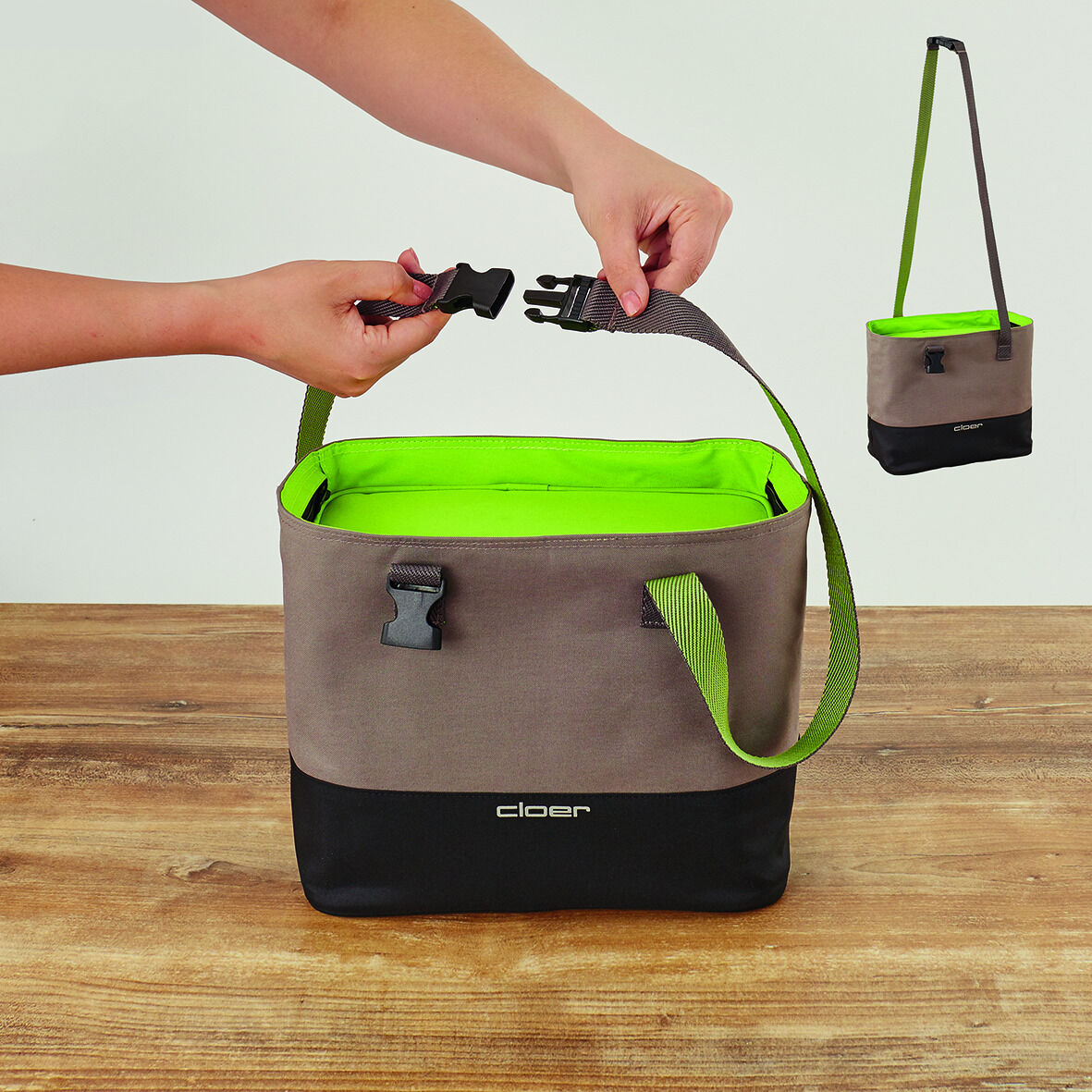 Lunch-Tasche in Beige und Grau mit grünem Innenfutter und Gurt wird von einer Hand gehalten. Auf einem Holztisch platziert. Eine kleinere Version der gleichen Tasche hängt an einem langen Gurt im Hintergrund.