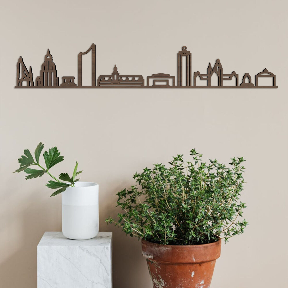 Holzschnitt der Skyline von Leipzig an einer beigen Wand hängend. Darunter steht ein weißer Marmorblock mit einer Vase mit grünen Blättern darauf, und daneben ein brauner Blumentopf mit einer Pflanze.