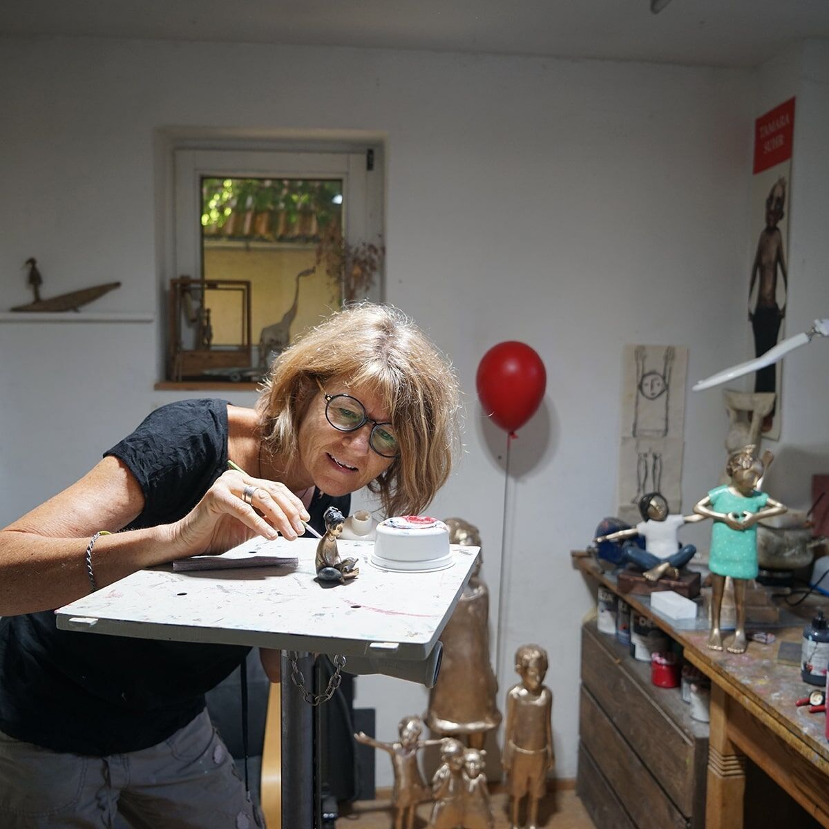 Eine Frau in einem Kunststudio arbeitet an einer Skulptur. Im Hintergrund sind weitere Kunstwerke und ein roter Luftballon zu sehen. Die Szene ist in natürlichen Farben gehalten.