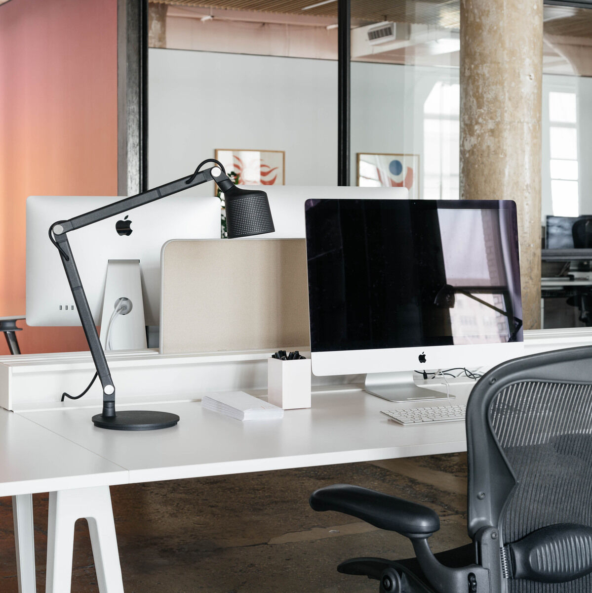 Eine Vipp Schreibtischlampe auf einem weißem Schreibtisch in einem modernen Büro, mit Computer und Bürostuhl im Hintergrund.