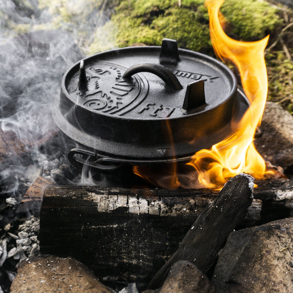 Ein Dutch Oven aus Gusseisen steht auf einer Feuerstelle im Freien, umgeben von Steinen und brennendem Holz, mit sichtbarem Rauch und Flammen im Hintergrund.
