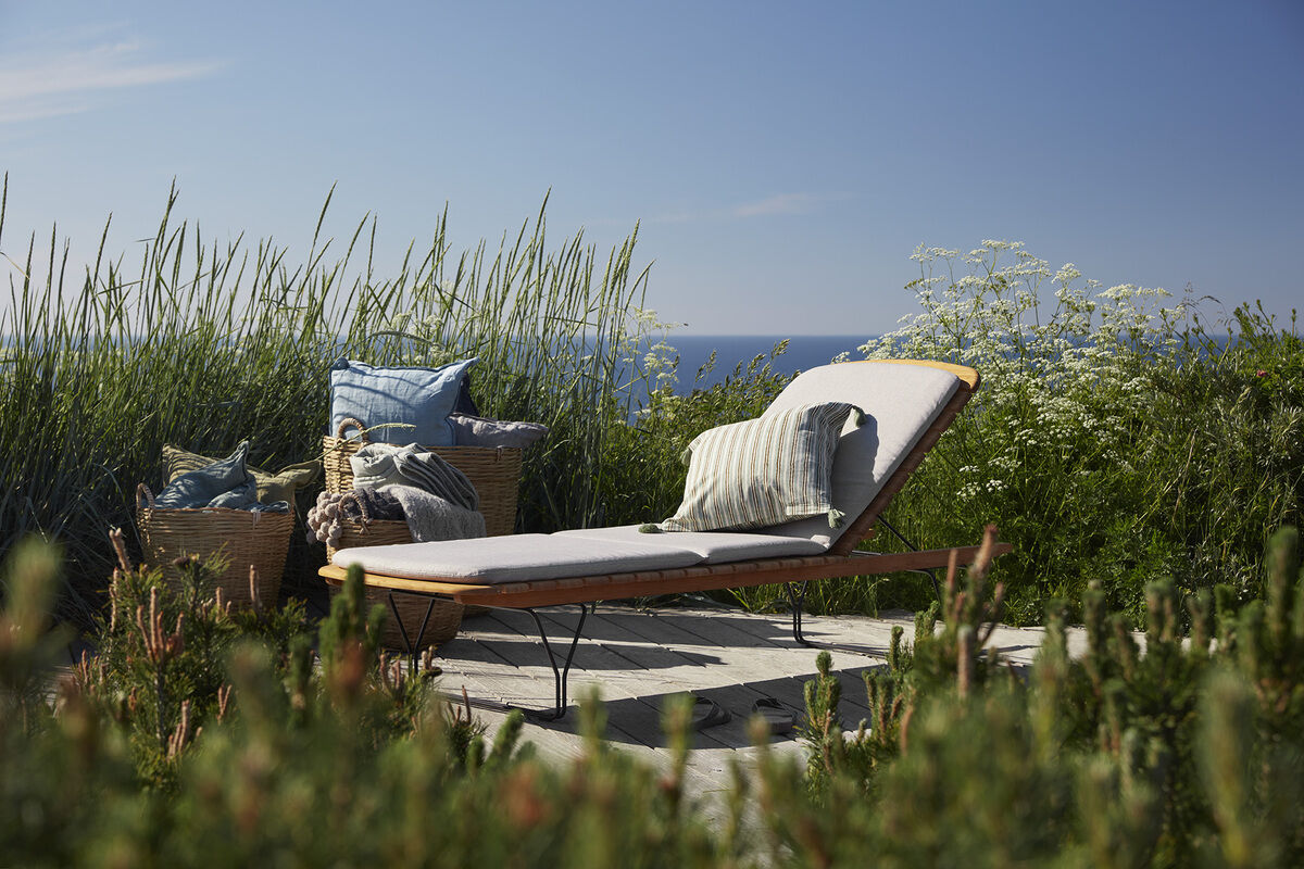 Ein gemütliches, hellblau-beiges Polsterkissen namens 'Auflage »MOLO«' auf einer Sonnenliege im Freien, umgeben von grüner Natur und einer malerischen Landschaft mit Blick auf das Meer im Hintergrund.