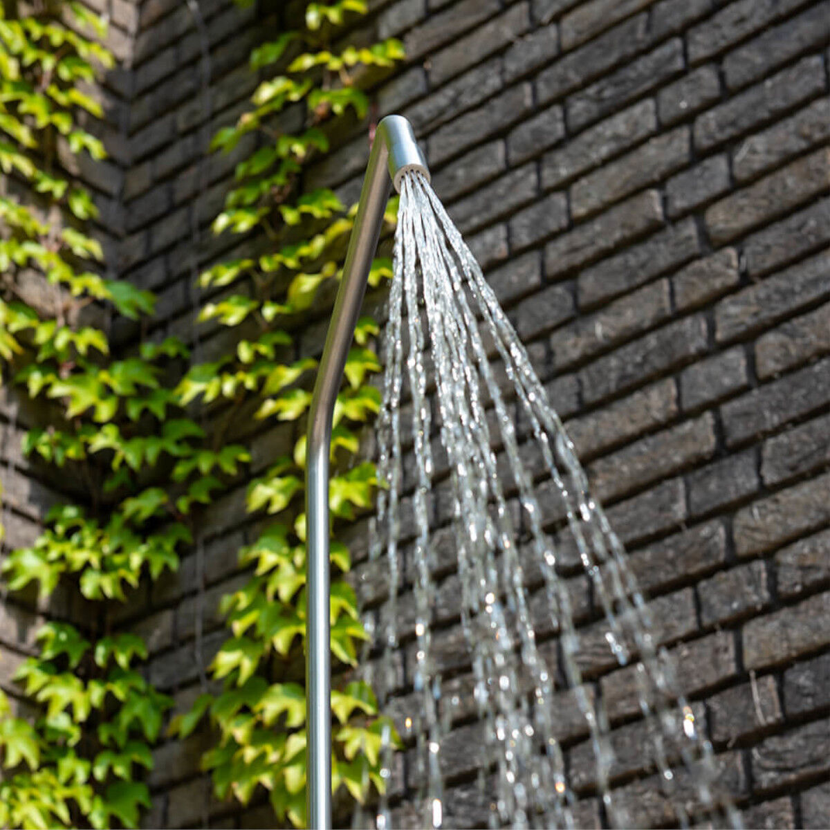 Die Gartendusche »Serpentine« von Weltevree steht vor einer gemauerten Wand mit Efeu. Aus dem geschwungenen, schlanken Edelstahlrohr fließt ein breiter, gleichmäßiger Wasserstrahl. Das Design ist minimalistisch und modern, die Szene wirkt erfrischend und stilvoll.
