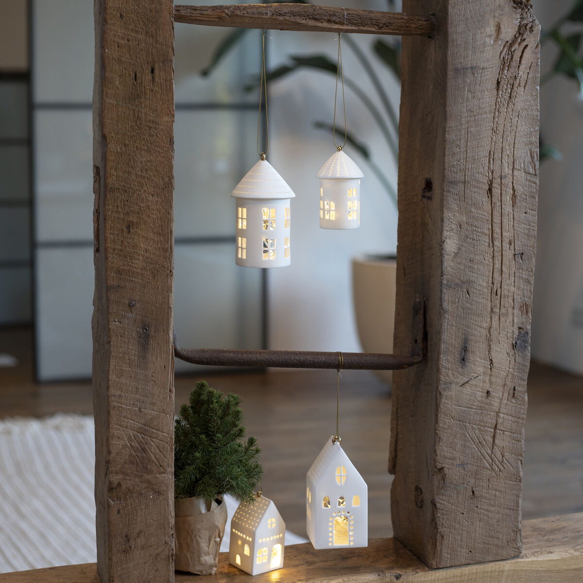 Hängende und stehende weiße Lichthäuser, beleuchtet von innen, im Holzrahmen einer rustikalen Leiter dekoriert. Ein kleiner Tannenbaum steht daneben.