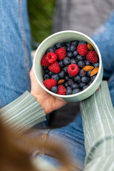 Person hält eine grüne Lunchbox mit Blaubeeren, Himbeeren und Mandeln, die auf ihrem Schoß sitzt.